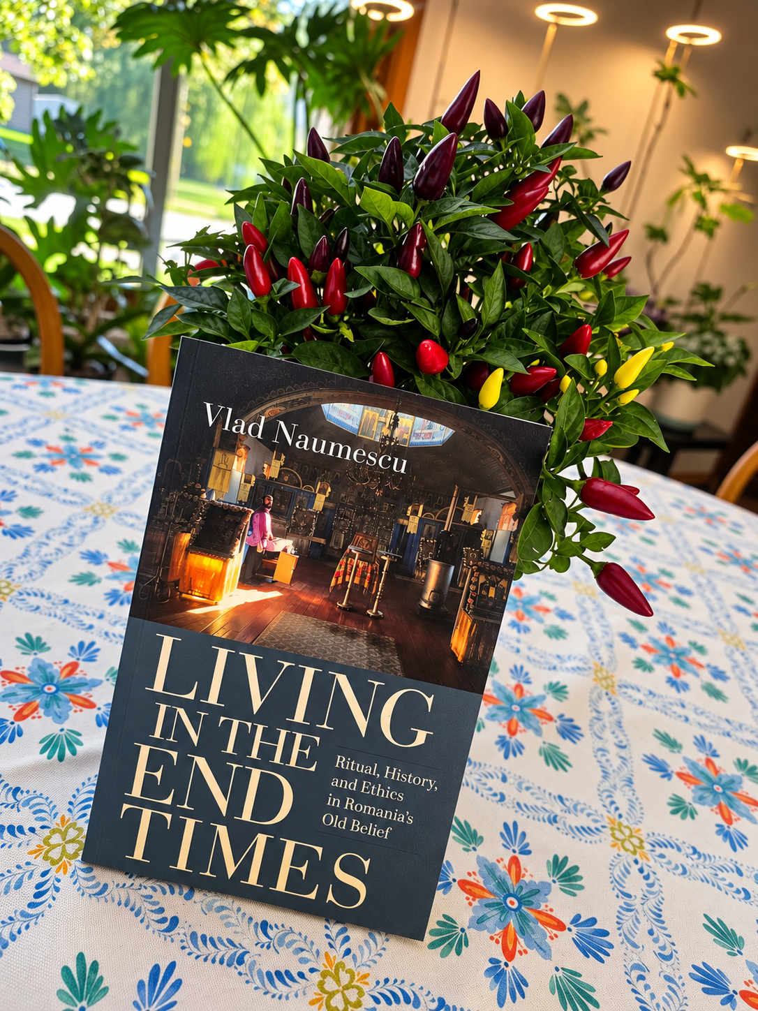 Living in the End Times by Vlad Naumescu beside a colorful pepper plant on a patterned tablecloth.